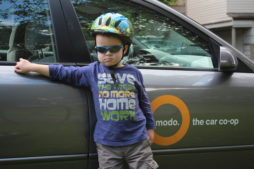 A child wearing a bike helmet stands in front of a Modo shared car